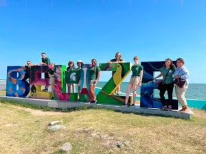 Photo of the MSU team posing on a colorful concrete sign that spells out 'BELIZE' on a sandy beach with a clear blue sky and the ocean in the background.