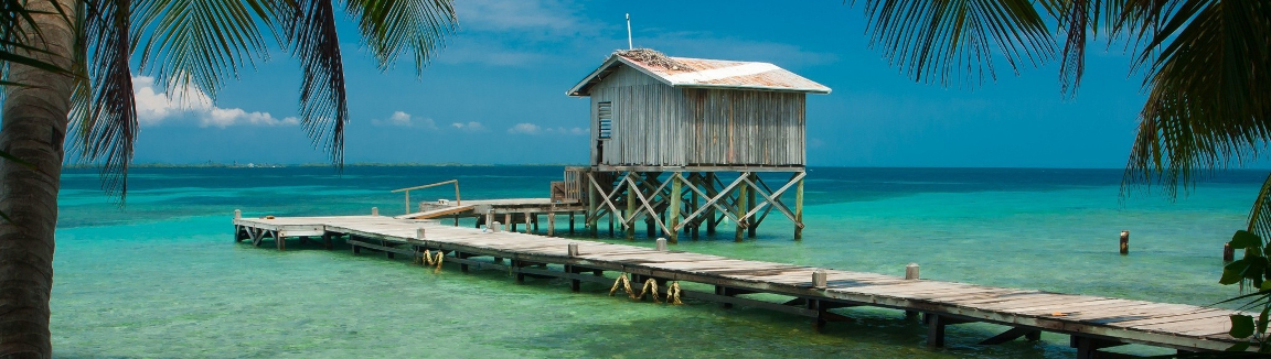 Photo of a weathered cabin on a dock on a beach in Belize. Palm trees frame the photo.